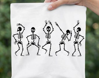 May include: A white t-shirt featuring a black graphic print of five skeletons in various dancing poses. The skeletons are arranged in a horizontal row, each displaying distinct arm and leg positions, creating a playful and rhythmic visual sequence across the fabric.