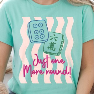 May include: A light teal t-shirt with a graphic of two Mahjong tiles and the text "Just one More round!" in pink script. The background has a wavy pattern of light pink and white stripes.