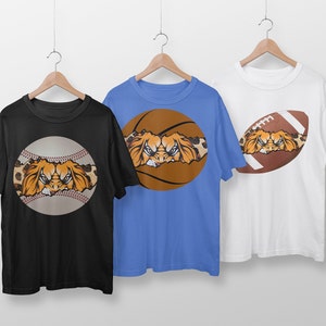 May include: Three t-shirts with different sports ball designs. Each shirt features a lion's face peeking out from behind a baseball, basketball, or football. The shirts are black, blue, and white.