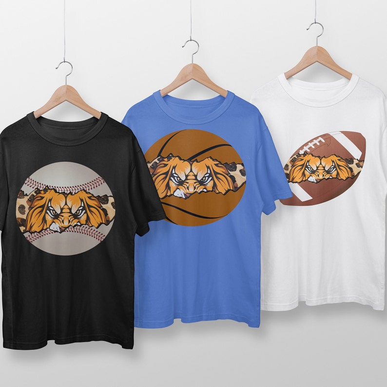 May include: Three t-shirts with different sports ball designs. Each shirt features a lion's face peeking out from behind a baseball, basketball, or football. The shirts are black, blue, and white.