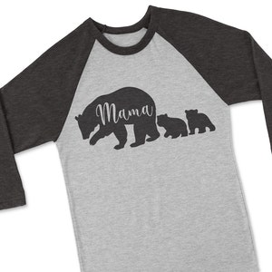 Puede incluir: Una camiseta de b&eacute;isbol gris y negra con una silueta de una osa madre y dos cachorros. La osa madre tiene la palabra "Mama" escrita en su espalda.