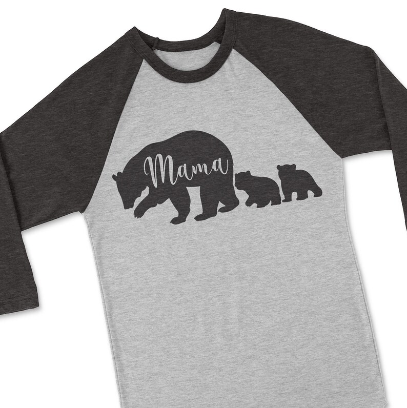 Puede incluir: Una camiseta de b&eacute;isbol gris y negra con una silueta de una osa madre y dos cachorros. La osa madre tiene la palabra "Mama" escrita en su espalda.