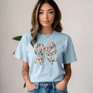 Pu&ograve; includere: Una maglietta blu chiaro con un design a fiocco floreale. Il fiocco &egrave; fatto di fiori colorati e ha un contorno dorato.