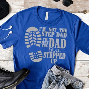 May include: A royal blue t-shirt with a grey print that reads "I'm not the step dad I'm just the dad that stepped up". The print features a large footprint and three smaller footprints.