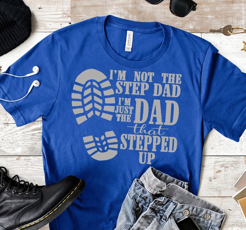 May include: A royal blue t-shirt with a grey print that reads "I'm not the step dad I'm just the dad that stepped up". The print features a large footprint and three smaller footprints.