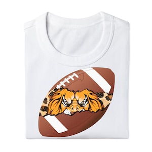 May include: A white t-shirt with a brown and white football graphic. The football is cracked open and a lion's face is visible inside. The lion's face is orange and brown with a black nose and eyes. The football is surrounded by a leopard print pattern.