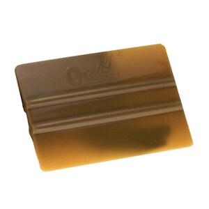 Puede incluir: Una espátula rectangular de color ámbar con el texto "OK" y un diseño de hoja. La espátula tiene crestas horizontales y probablemente se utiliza para alisar superficies o aplicar materiales.