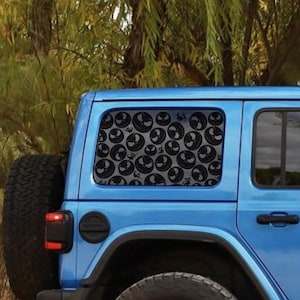 May include: A blue Jeep with a rear window covered in a pattern of black Jack Skellington faces. The faces are repeated across the window, creating a unique design. The vehicle is parked outdoors with trees in the background.