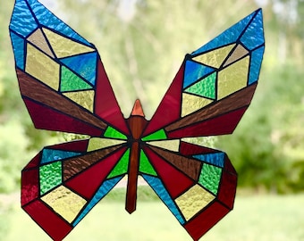 May include: A stained glass butterfly with a geometric design. The wings feature sections of blue, green, gold, and brown glass, outlined in red. The butterfly's body is brown with a red center, and it is set against a blurred green background.