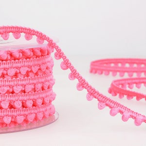 Puede incluir: Borde de pompones rosa con un carrete de borde. El borde tiene pequeños pompones redondos a lo largo del borde.