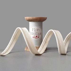 Puede incluir: Una bobina de cinta de bies de doble pliegue de color blanco roto con una bobina de madera y una etiqueta que dice "Plum Boutique".