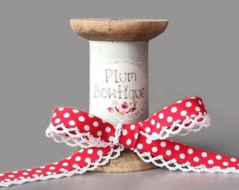 Könnte beinhalten: Rot-weißes gepunktetes Band mit weißer Spitzenborte, das um eine Holzspule gewickelt ist. Die Spule trägt ein Etikett mit der Aufschrift "Plum Bowtique".