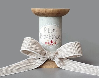 May include: A spool of beige ribbon tied with a bow. The spool is wooden and has a blue and white label that says "Plum Boutique".