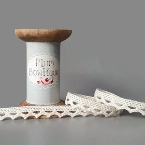 May include: A light blue thread spool with a label reading "Plum Boutique", and a length of off-white lace with a scalloped edge. The spool is made of wood and cardboard.