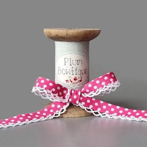 Könnte beinhalten: Rosa-weißes gepunktetes Band mit weißer Spitzenborte, um eine Holzspule gewickelt. Die Spule trägt ein Etikett mit der Aufschrift "Plum Bowtique".
