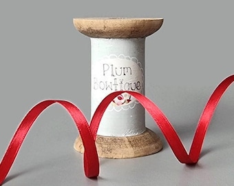 Puede incluir: Una bobina de madera con una cinta de raso roja. La bobina tiene una etiqueta azul claro con el texto "Plum Boutique". La cinta roja vibrante se curva a través de la imagen.
