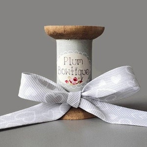 Puede incluir: Un carrete de madera con una cinta de rayas blancas y azules claras envuelta alrededor. El carrete está decorado con una etiqueta que dice "Plum Boutique" con un diseño floral.