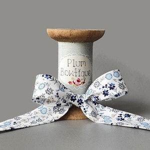 Può includere: Un rotolo di nastro bianco con un motivo floreale blu. Il rotolo è in legno e ha un'etichetta blu con la scritta "Plum Boutique".