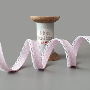 Könnte beinhalten: Rosa-weißes gepunktetes Band mit weißer Spitzenborte. Das Band ist um eine Holzspule gewickelt, die mit "Plum Boutique" beschriftet ist.