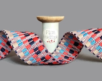 May include: A woven fabric ribbon featuring a multi-colored pattern of red, navy blue, light blue, and cream rectangular blocks. The ribbon is wrapped around a wooden spool with a label that reads Plum Boutique in black text.