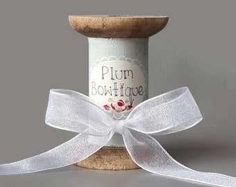 May include: A wooden spool with a light blue label that reads "Plum Bowtique" is adorned with a white ribbon tied in a bow. The spool has a light brown top and bottom. The ribbon is a sheer, shimmering white.