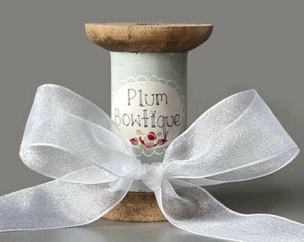 May include: A wooden spool with a light blue and white label that says "Plum Boutique" with a floral design. A white sheer ribbon is tied around the spool.