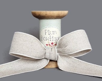 May include: A light beige ribbon tied in a bow, wrapped around a wooden spool. The spool has a light blue label with the text "Plum Boutique" and a floral design. The ribbon has a textured appearance and a white border.