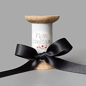 Puede incluir: Un carrete de madera con una cinta de satén negra atada en un lazo. El carrete está envuelto en papel blanco con una etiqueta que dice "Plum Boutique".