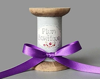 May include: A wooden spool painted light grey with a label that reads "Plum Boutique". A purple satin ribbon is tied around the spool in a bow.