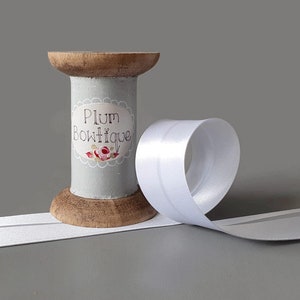 Puede incluir: Una bobina de cinta de satén blanco con una base de madera. La bobina está decorada con una etiqueta que dice "Plum Boutique".