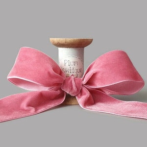Peut inclure: Un ruban de velours rose noué en un nœud sur une bobine en bois. La bobine porte une étiquette sur laquelle est inscrit "Plum Boutique".