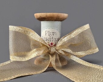 May include: A spool of gold ribbon with a bow tied around it. The spool is decorated with a blue and white label that says "Plum Boutique".