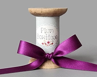 May include: A spool of purple satin ribbon tied into a bow, sitting on a wooden spool with a label that reads "Plum Boutique".