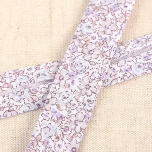 May include: Two strips of floral fabric with a repeating pattern of small, light purple and brown flowers on a white background. The fabric is folded over, creating a double-layered effect. The background is a neutral beige.