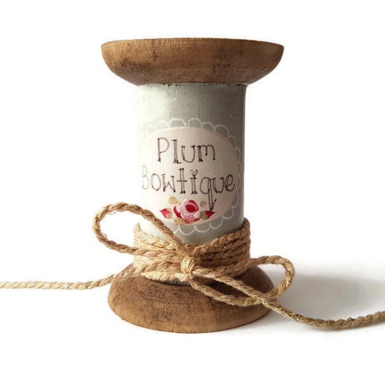May include: A wooden spool painted light grey with a label that reads "Plum Boutique" and a floral design. The spool is wrapped with twine and tied with a bow.