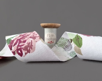 May include: White cotton ribbon with a floral print of pink and burgundy roses. The ribbon is wrapped around a wooden spool with a label that reads "Plum Bowtique".