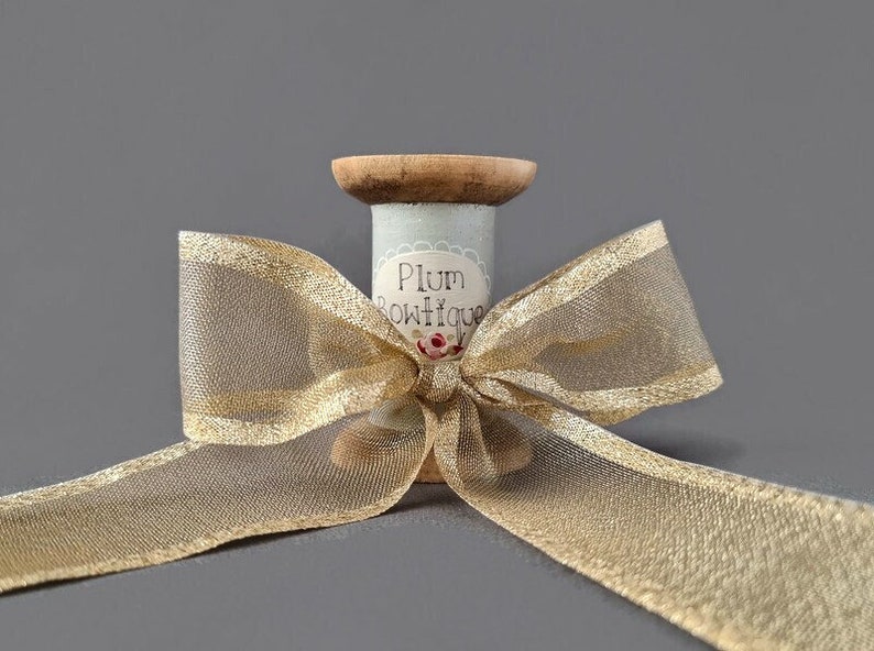 May include: A spool of gold ribbon with a bow tied around it. The spool is decorated with a blue and white label that says "Plum Boutique".