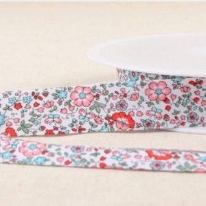Puede incluir: Cinta de algodón blanca con estampado floral en tonos rosa, rojo y azul. La cinta está en un carrete blanco.