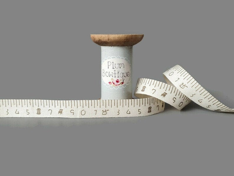 Puede incluir: Una bobina de cinta de tela blanca con una bobina de madera marr&oacute;n y una etiqueta que dice "Plum Boutique". La cinta es una cinta m&eacute;trica con n&uacute;meros y marcas marrones.