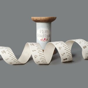 Puede incluir: Una cinta de algod&oacute;n blanca con medidas impresas en marr&oacute;n y un icono de aguja de coser. La cinta est&aacute; enrollada en un carrete de madera con una etiqueta que dice "Plum Boutique".