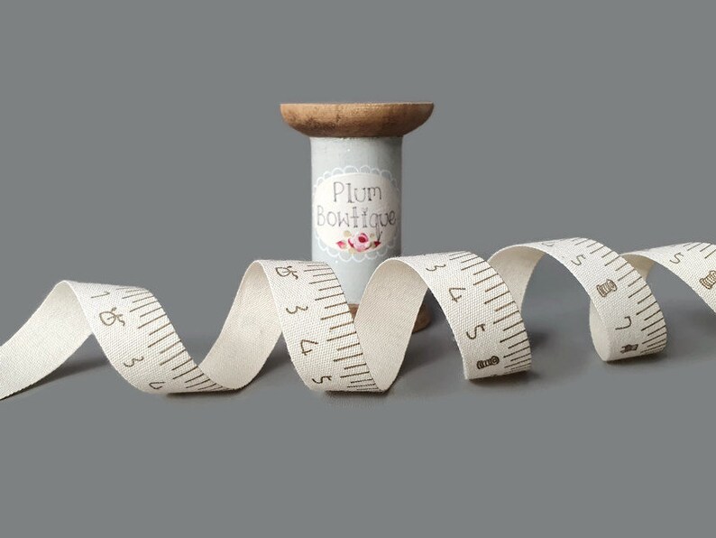 Puede incluir: Una cinta de algod&oacute;n blanca con medidas impresas en marr&oacute;n y un icono de aguja de coser. La cinta est&aacute; enrollada en un carrete de madera con una etiqueta que dice "Plum Boutique".