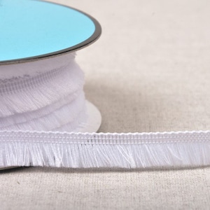 May include: White fringe trim with a delicate texture. The trim is wound around a spool with a light blue label.