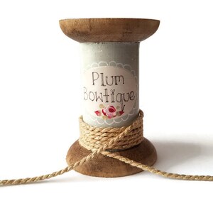 May include: A wooden spool painted light blue with a label that reads "Plum Boutique" and a floral design. The spool is wrapped with a gold and natural coloured twine.