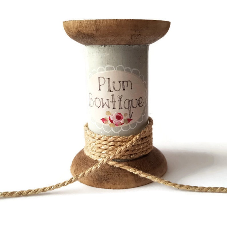 May include: A wooden spool painted light blue with a label that reads "Plum Boutique" and a floral design. The spool is wrapped with a gold and natural coloured twine.