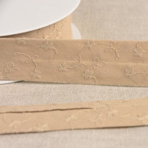Könnte beinhalten: Beigefarbenes Baumwollband mit einem zarten floralen Stickmuster. Das Band ist in der Mitte gefaltet, so dass die Nähte auf der Rückseite sichtbar sind.