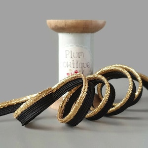 Könnte beinhalten: Schwarzes und goldenes elastisches Band mit einem goldenen Metallic-Faden entlang der Kante. Das Band ist um eine Holzspule mit einem Etikett mit der Aufschrift "Plum Boutique" gewickelt.