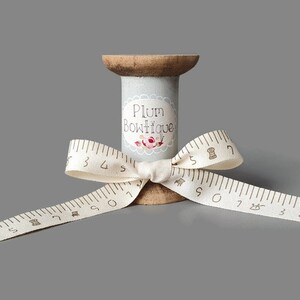 Puede incluir: Un carrete de madera con una etiqueta gris que dice "Plum Boutique" y un dise&ntilde;o floral. Una cinta blanca con marcas marrones est&aacute; atada alrededor del carrete en forma de lazo.