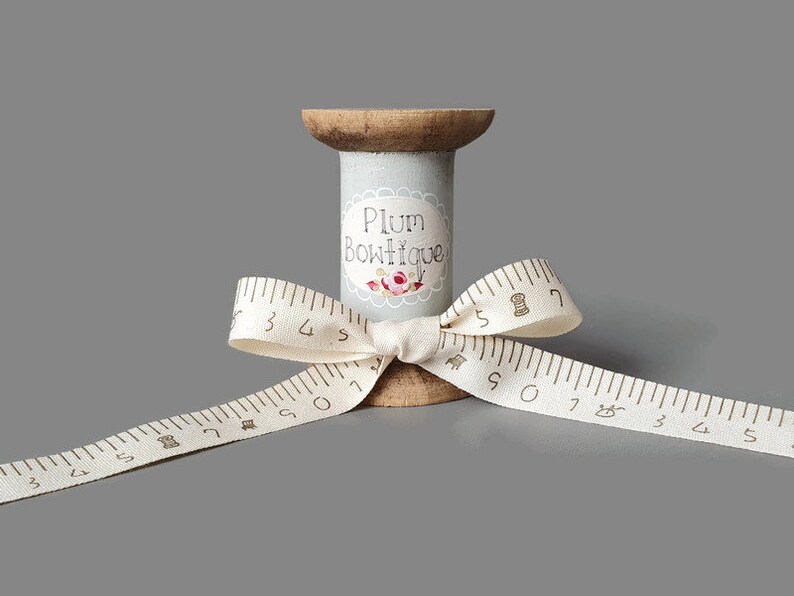 Puede incluir: Un carrete de madera con una etiqueta gris que dice "Plum Boutique" y un dise&ntilde;o floral. Una cinta blanca con marcas marrones est&aacute; atada alrededor del carrete en forma de lazo.