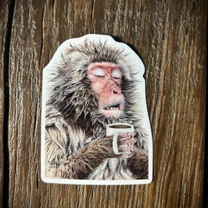 May include: A close-up of a snow monkey with its eyes closed, holding a white mug with a brown liquid inside. The monkey has a brown and white fur coat and is sitting on a white background.