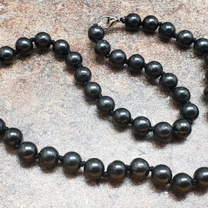 May include: A black beaded necklace with a silver clasp. The necklace features evenly spaced, round, dark gray beads strung on a black cord. The beads have a glossy finish. The necklace is displayed on a textured, neutral-toned surface.